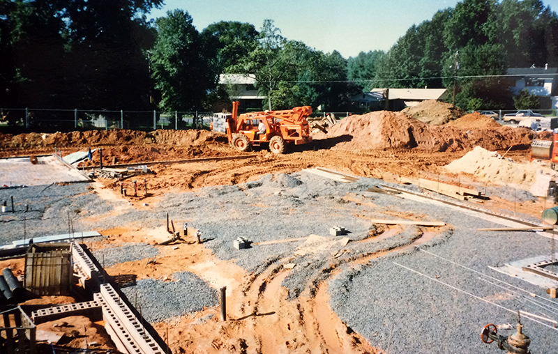 Undated color photograph showing construction progress on a new addition to Rolling Valley Elementary School. The foundation has been outlined with cinderblock. Gravel has been placed, preparing the ground for the concrete pad. In the distance, a forklift is moving cinderblocks. 