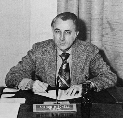 Black and white yearbook portrait of Principal Arthur Mitchell taken in 1973. He is seated at his desk signing papers.